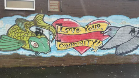 A spray painted green fish, grey pigeon and red love heart with the words "love your community" on a yellow spray painted ribbon.