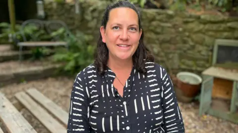The picture shows a person sitting outdoors at a wooden picnic-style table in a garden or courtyard area, wearing a dark dress with a white abstract pattern and a small microphone clipped to the front of the clothing. The background includes a stone wall, gravel ground, plants and trees, a metal bench, and some weathered outdoor furniture.