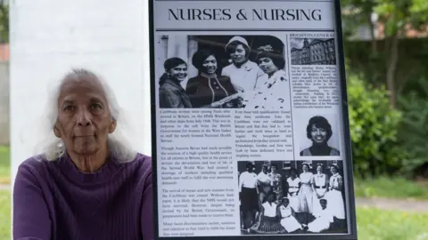 JJ Waller/Brighton & Hove Museums Shirley Williams sits next to a framed image titled "nurses & nursing," describing how migrants from the Caribbean and across the Commonwealth who came to Britain on the HMT Empire Windrush contributed to the National Health Service (NHS) post-World War II. It features black and white images of Shirley as a young woman.