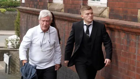 PA Ray and Campbell Hatton. Ray wears a light-coloured  quarter-zip with a glasses chain on and carries a jacket. Campbell is wearing a three-piece black suit, with white shirt, black tie and silver tie clip. They are walking on a pavement next to a red-brick building.