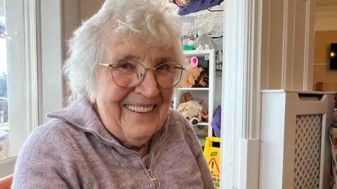 Patricia, a resident at the care home, with a big smile as she looks at the camera. Patricia is wearing glasses and has a lilac fleece zip top on. Behind her, there are some shelves with toys on and a doorway.