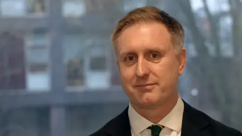 Vikki Irwin/BBC A close-up of Ed Garratt wearing a white shirt with tie and black suit jacket over the top. He is looking directly into the camera