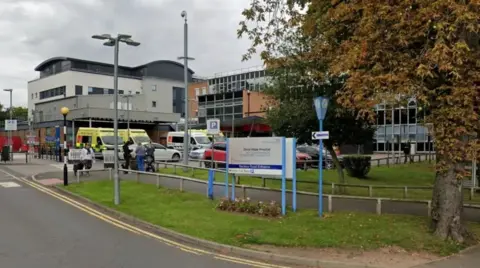 The entrance to a hospital site with a road running on front of large multi storey buildings and a blue NHS sign