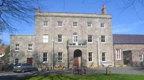 A three story building made out of grey stone, with a lawn in front.