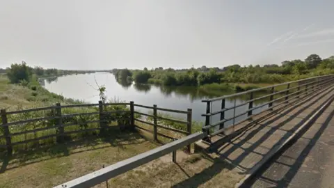 Google A view of the Jubilee River from a bridge, on a sunny day. It is quite a wide river with fields and bushes on either side.