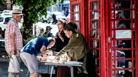 Adam Victor Lisa Wilson and Kim Squirrell standing behind a table next to the two red phone boxes. They are speaking to two men. One of them is bending over the table and writing.