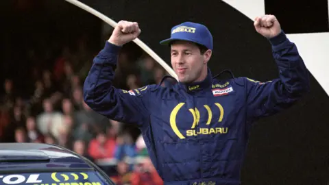 PA Media Colin McRae in a blue boiler suit with Subaru branding. He also has a blue Pirelli baseball cap. His arms are in the air in celebration. He's standing next to a rally car, in front of a crowd of people