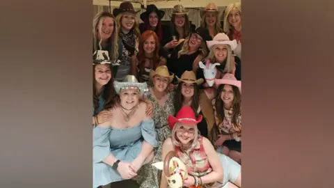Robyn Law A group of around 15 women wearing cowboy hats smile for the camera at a Western-themed party. Most of them have wigs but some are bald. A couple of them have inflatable hobby horses.