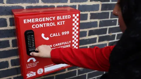 A woman trying to access the Cowley bleed kit. She is wearing a red long-sleeve top. He face is visible only in profile and she is wearing a black hijab.