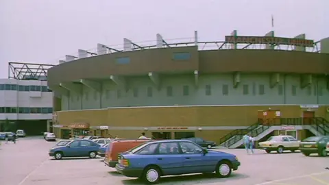 Cars parked by the side of Old Trafford underneath a red and black Manchester United sign in 1989