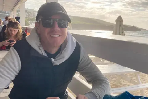 Patrick Charnley Patrick smiling and sitting at a beachfront bar, wearing sunglasses