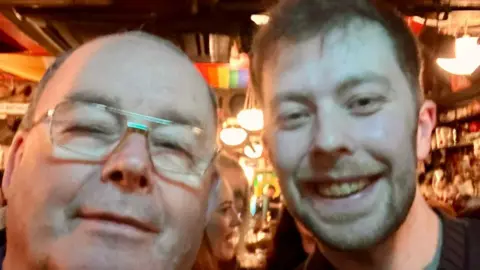 Dougie Breslin Two men appear close to camera in a selfie shot. The man on the leftis bald and wear glasses, while the man on the right has short brown hair and is smiling. They are standing in a pub.