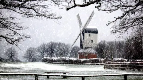 Dr Edmund Ranasinghe Stock Windmill in a snowstorm. It shows a windmill with red brick base, and a wooden structure with two white sails. There are trees behind the windmill, bushes in front, and a short fence.