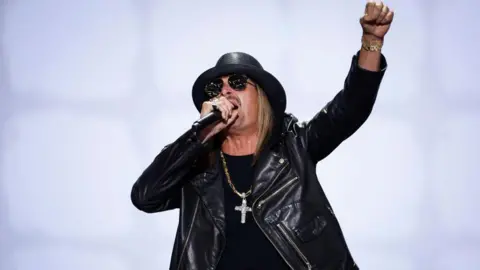 Musician Kid Rock sings into a microphone and lifts a fist. He is wearing a black leather jacket, T-shirt, hat and sunglasses and a large silver cross on a chain.