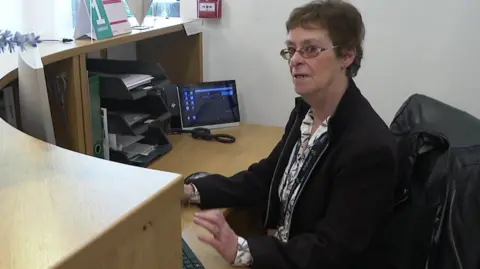 A woman with short dark hair and glasses is sat behind a desk, looking to the camera. There is a phone and a computer on the desk and she is wearing a black jacket with a white floral shirt.