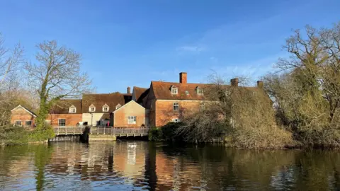 Babergh District Council A series of red brick old buildings beside, and reflected in, the river Stour in the foreground. Part of a building to the right of the image is obscured by a large overhanging tree, part of which is trailing in the water. 