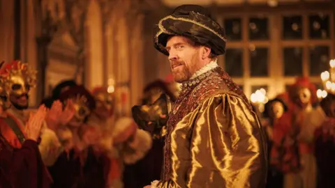 Actor Damian Lewis as King Henry VIII in Wolf Hall. The scene is warmly lit and he is surrounded by people wearing masquerade-type masks and applauding. He is wearing a large black hat, a shiny gold-coloured top with puffy sleeves and an elaborately decorated waistcoat. He is standing side on to the viewer but looking back towards the camera with a smirk.