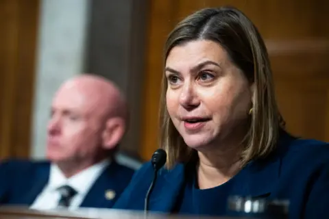 Michigan Senator Elissa Slotkin appears in Congress speaking into a microphone while Arizona Senator sits behind her out of focus.