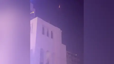 Teigan McFadden A photo with a purple hue of an intercepted missile. It can be seen falling to earth as a small orange streak in the night's sky. Below it a two-storey Middle Eastern style building can be seen with a white facade.