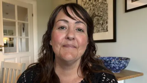 Kelly Davison has shoulder-length, dark hair. She is sitting at a kitchen table and smiling at the camera. She is wearing a black top. There are pictures behind her and a door off to the left. 