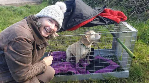 Maxine Young is crouching down to a crate which has a small pale labrador puppy in it. Ms Young is smiling and wearing a brown coat and woolly hat. The puppy is sitting on a fleece in the crate which is lying on grass. 