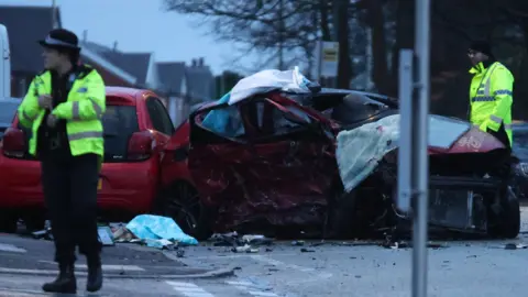Crash scene shows two badly damaged cars and debris in the road. Two police officers wearing yellow hi-vis jackets are at the scene.