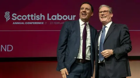 PA Media Anas Sarwar, with black hair, stands arm in arm in Keir Starmer, with grey hair and glasses, on a stage. Both are wearing dark suits and smiling in front of a Scottish Labour banner 