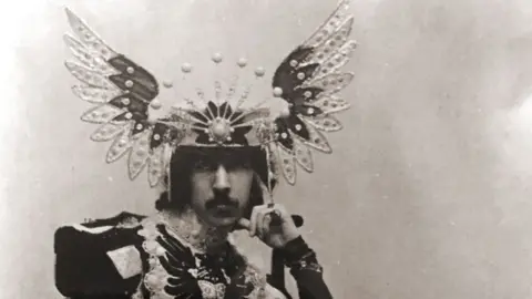 The 5th Marquess of Anglesey in a sepia photograph wearing a floor length overcoat consisting of differently coloured pieces of heavy material. The top of the coat is military in style with large shoulder pieces. He wears a headdress of splayed wings with a tiara-effect frontspiece.