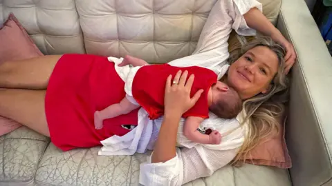 A woman with long blonde hair, wearing a white top and red skirt lying on a cream couch, with a baby sleeping on her chest.