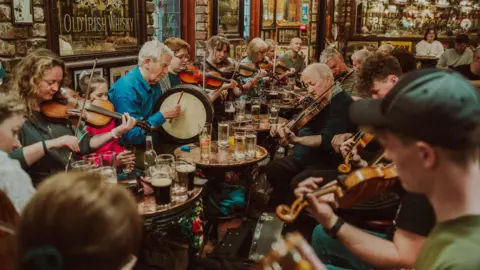 TradFest People sitting in a pub are playing instruments. Many are playing the fiddle. The pub is crowded. Drinks are sitting on tables. 