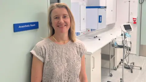 Dr Isabel Smith posing next to the open pale-green door to the anaesthetic room. Behind her is a white counter top, a drip trolley and a heart monitor on another trolley stand. She has shoulder length blonde hair and is wearing a grey, lace-patterned, short-sleeved top.