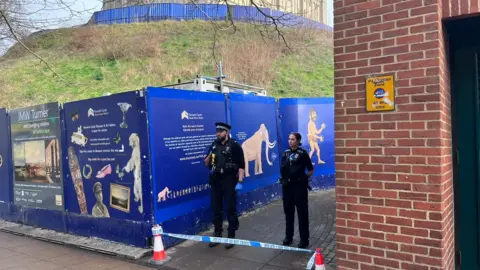 Neve Gordon-Farleigh/BBC Two police officers stand at a police cordon area. The cordon is at a walkway of Norwich Castle and in front of the officers are two cones with blue and white police tape on.