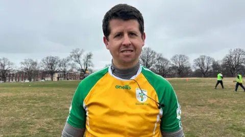 Kevin Tobin, he has short black hair and is wearing a GAA jersey which has a yellow trunk and green sleeves over a grey long-sleeved top. He is standing in a park.