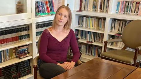 A girl in her 20's with blonde hair and blue eyes is sitting in a chair, looking off centre from the camera.. She is wearing a purple top and necklace and black jeans. In the background is a chair and bookcases.
