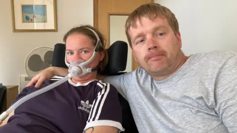 A woman is sitting down with a breathing apparatus on. She is wearing a blue Adidas t-shirt. Sitting with her is a man, wearing a grey t-shirt. 