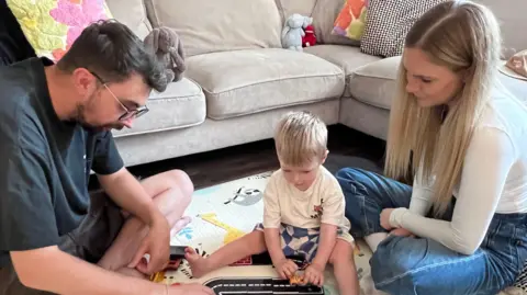 A blonde haired two year old sits playing with a toy car on the floor between his mother and father.