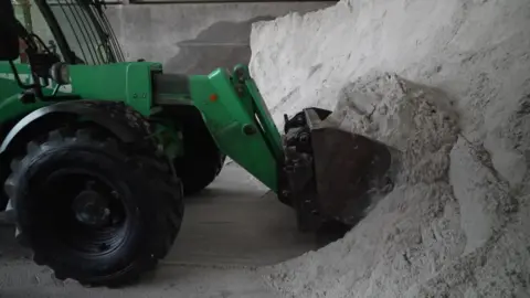 Peter Whittlesea/BBC A green tractor with a scoop on the front shovelling a pile of grey gritting salt.