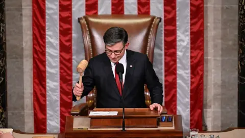 Mike Johnson wearing a black suit, red tie and slams a gavel in front of an American flag