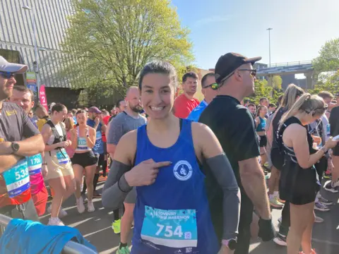 Isobel Lockley is wearing a blue vest and pointing to a badge on her chest. She is smiling and wearing a number badge for the Coventry half marathon