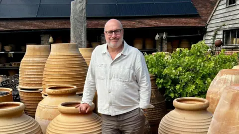 A bald man wearing a white shirt stood among lots of large ceramic pots outside. He is smiling at the camera and has his right hand on top of a pot and his left in his pocket