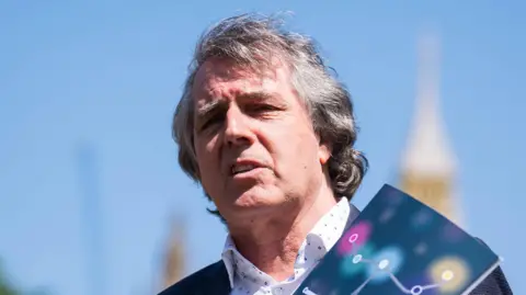PA Media Steve Rotheram speaking at a press call earlier this year in Westminster. He is holding a booklet up while talking. The backdrop is a clear blue sky with Houses of Westminster in the background