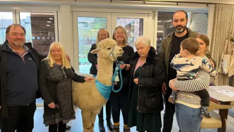 Barbara Wild, wearing a pink top and coat, stands next to Prancer the alpaca, who has a blue lead. There are other residents and staff around the pair.