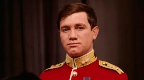 National Portrait Gallery A cropped version of a National Portrait Gallery image of army officer Captain Robert Nairac. He has short brown hair and wears a red military jacket decorated with gold buttons, gold epaulettes, and a single medal pinned to his chest. He is holding a large black bearskin hat.