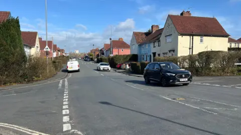 Guildford Road in Brislington. It has a number of shops, with cars parked on the pavement. Terraced residential properties are lined in the distance.