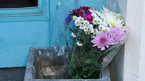 A bouquet of flowers, including white, pink, yellow, and deep purple blooms, rests inside a square stone planter placed beside a weathered turquoise door.