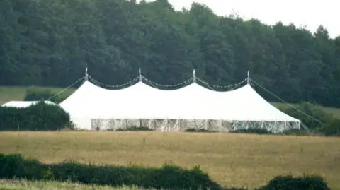 Steve Parsons/PA A view across a field showing four peaks of a large white marquee set up in the countryside. There are fairy lights draped in a string over the top and in the background there is a row of large, dense green trees.