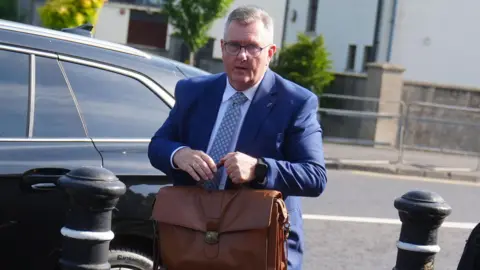 PA Media Sir Jeffrey Donaldson has short, cropped grey hair. He is wearing black-rimmed glasses and a blue suit, white shirt and blue patterned tie. He is standing in front of a black car and is carrying a brown leather satchel bag in front of him. 