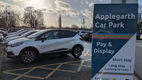 A car park with several parked vehicles and a large “Applegarth Car Park” sign showing pay‑and‑display information, with a CCTV warning sign above it in low afternoon light.