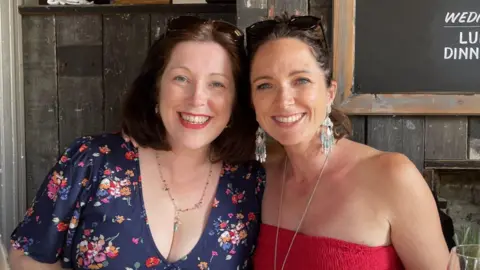 Jo O'Connell Lisa Mountain Thompson and Jo O'Connell smile at the camera, standing side-by-side. Both have shoulder-length brown hair with sunglasses pushed back over their heads and wear colourful summer clothes.