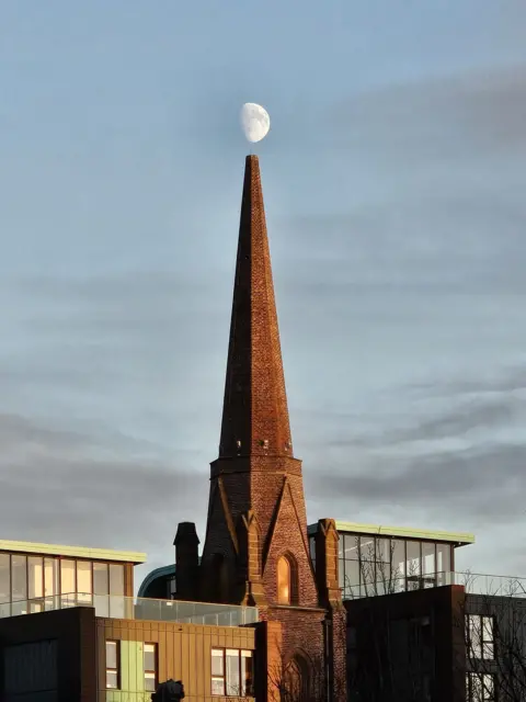 Neil Moir Una torre de iglesia de ladrillo rojo con la luna vista directamente encima de ella.